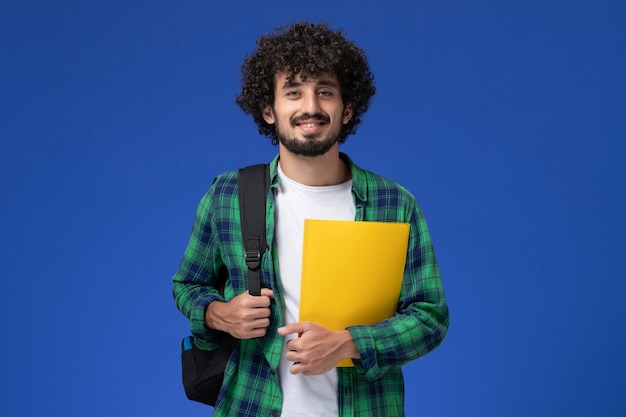 Vista frontal de um estudante do sexo masculino com camisa quadriculada verde, usando uma mochila preta e segurando arquivos na parede azul