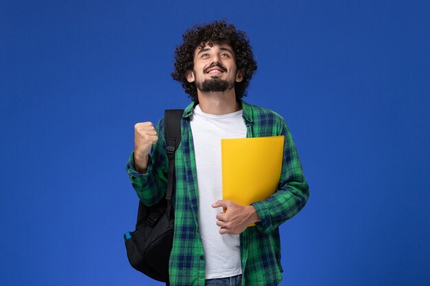 Vista frontal de um estudante do sexo masculino com camisa quadriculada verde, usando uma mochila preta e segurando arquivos na parede azul