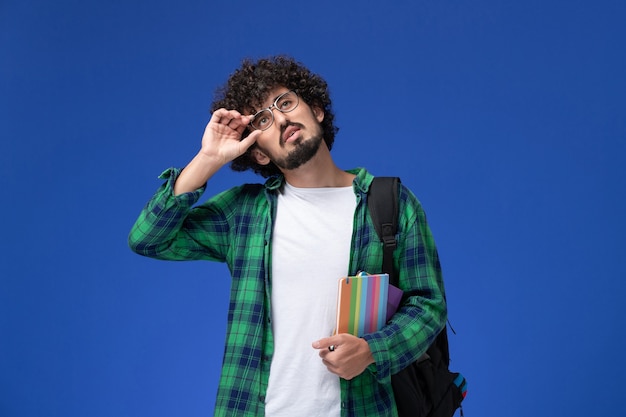 Vista frontal de um estudante do sexo masculino com camisa quadriculada verde, usando uma mochila preta e segurando o caderno na parede azul