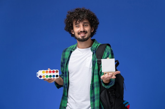 Vista frontal de um estudante do sexo masculino com camisa xadrez verde e mochila preta segurando pinturas e cavalete na parede azul