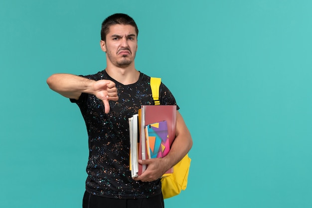 Vista frontal de um estudante do sexo masculino com camiseta escura e mochila amarela segurando o caderno e arquivos na parede azul