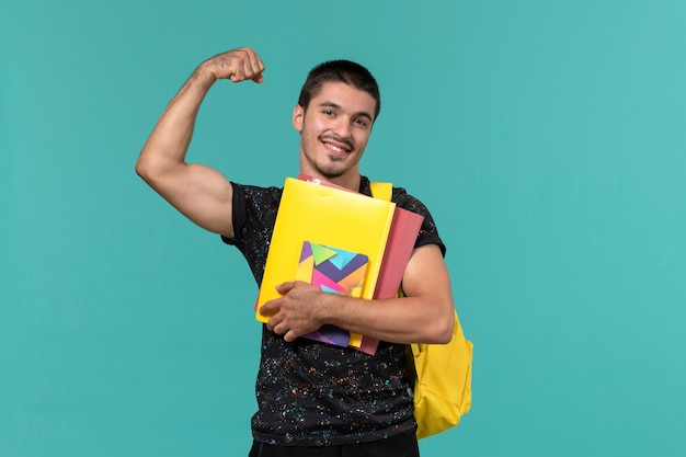 Vista frontal de um estudante do sexo masculino em uma mochila de camiseta amarela escura segurando arquivos e o caderno na parede azul clara