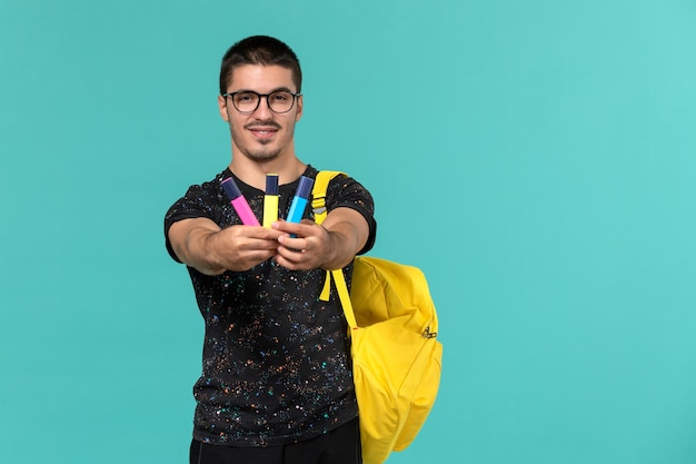 Vista frontal de um estudante do sexo masculino em uma mochila de camiseta amarela escura segurando canetas de feltro coloridas na parede azul