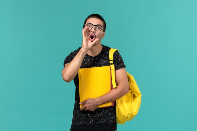 Vista frontal de um estudante do sexo masculino em uma mochila de camiseta amarela escura segurando diferentes arquivos chamando na parede azul claro