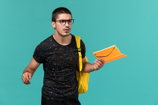 Vista frontal de um estudante do sexo masculino em uma mochila de camiseta amarela escura segurando o caderno na parede azul