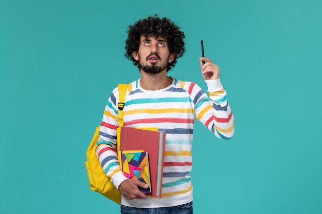 Vista frontal de um estudante do sexo masculino usando uma mochila amarela segurando uma caneta e um caderno de arquivos na parede azul