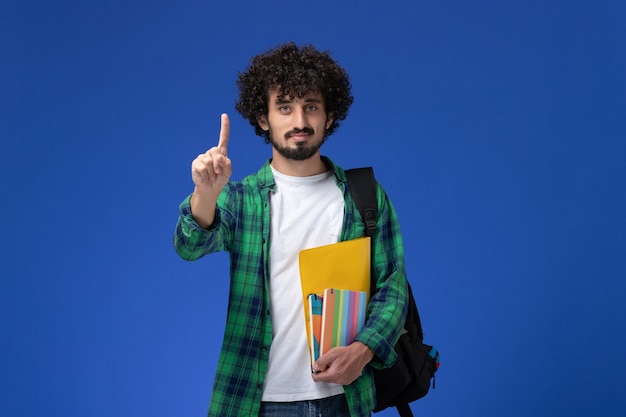 Vista frontal de um estudante do sexo masculino usando uma mochila preta segurando cadernos e arquivos na parede azul