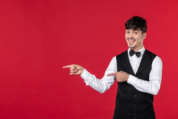 Vista frontal de um garçom sorridente de uniforme com gravata borboleta no pescoço e apontando algo do lado direito na parede vermelha