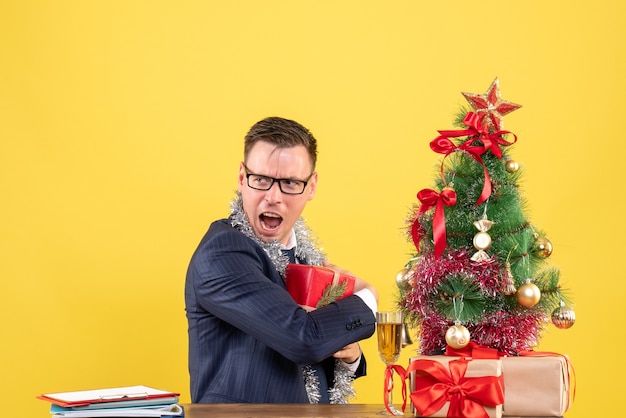 Vista frontal de um homem agitado, escondendo seu presente, sentado à mesa perto da árvore de natal e presentes na parede amarela