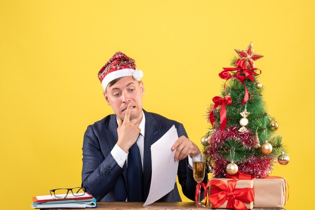 Vista frontal de um homem agitado sentado à mesa perto da árvore de natal e presentes na parede amarela