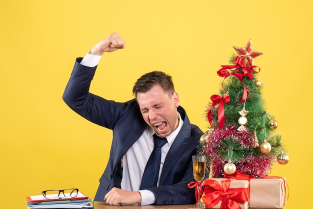 Vista frontal de um homem bonito, mostrando o gesto vencedor, sentado à mesa perto da árvore de natal e presentes em amarelo