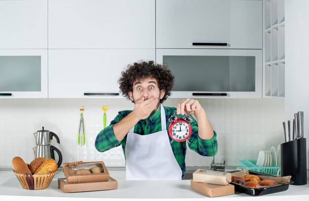 Foto grátis vista frontal de um homem chocado em pé atrás da mesa com vários doces e segurando o relógio na cozinha branca