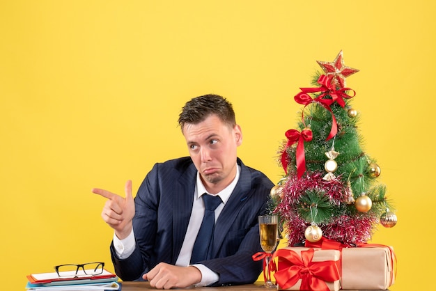 Vista frontal de um homem confuso apontando algo sentado à mesa perto da árvore de natal e presentes em amarelo