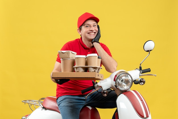 Vista frontal de um homem de correio esperançoso, sorridente, usando uma blusa vermelha e luvas de chapéu com máscara médica, entregando pedidos sentado em uma scooter segurando pedidos