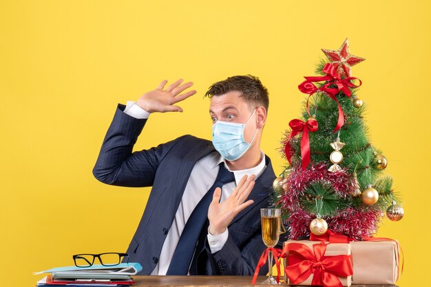 Vista frontal de um homem de negócios assustado abrindo as mãos, sentado à mesa perto da árvore de natal e presentes em amarelo