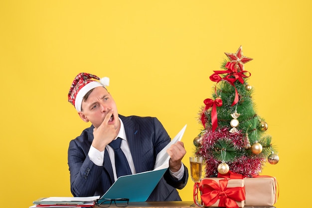 Vista frontal de um homem de negócios confuso sentado à mesa perto da árvore de natal e presentes em amarelo