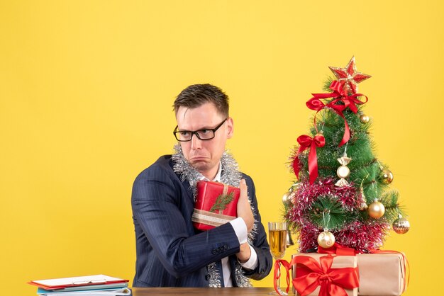 Vista frontal de um homem deprimido, escondendo seu presente, sentado à mesa perto da árvore de natal e presentes em amarelo