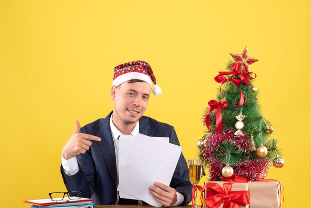 Vista frontal de um homem feliz apontando para documentos sentados à mesa perto da árvore de natal e presentes em amarelo