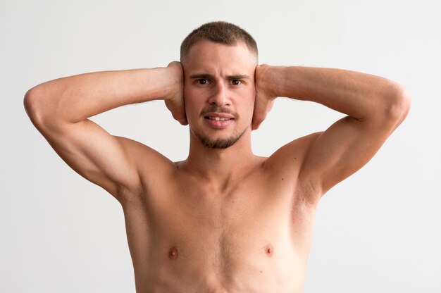 Vista frontal de um homem sem camisa exibindo seus bíceps enquanto cobre as orelhas