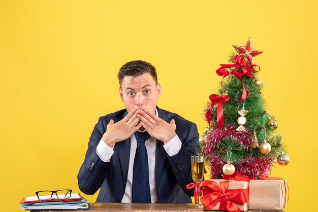 Vista frontal de um homem surpreso colocando as mãos na boca, sentado à mesa perto da árvore de natal e presentes na parede amarela