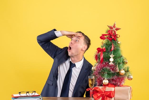 Vista frontal de um homem surpreso olhando algo sentado à mesa perto da árvore de natal e presentes na parede amarela