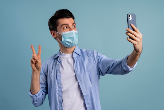 Vista frontal de um homem usando máscara médica e tirando uma selfie enquanto faz o sinal da paz