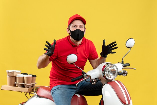 Vista frontal de um jovem adulto curioso vestindo blusa vermelha e luvas de chapéu na máscara médica, entregando o pedido sentado na scooter sobre fundo amarelo