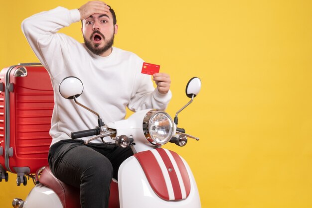 Vista frontal de um jovem chocado viajando sentado em uma motocicleta com uma mala segurando um cartão de banco em fundo amarelo isolado