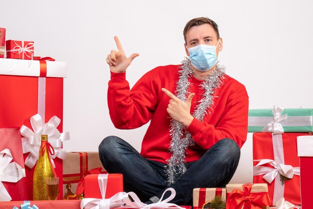 Foto grátis vista frontal de um jovem com máscara sentado ao redor do natal apresenta-se na parede branca