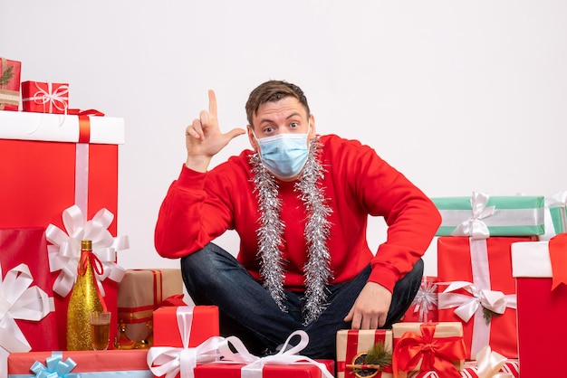 Foto grátis vista frontal de um jovem com máscara sentado ao redor do natal apresenta-se na parede branca