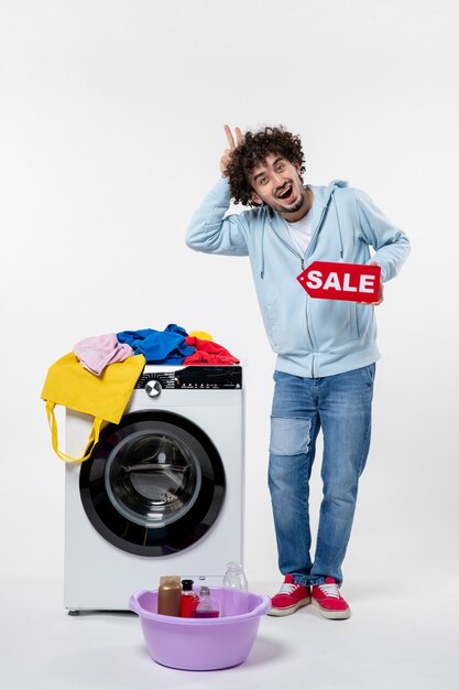 Vista frontal de um jovem com uma lavadora segurando uma bandeira vermelha de venda na parede branca