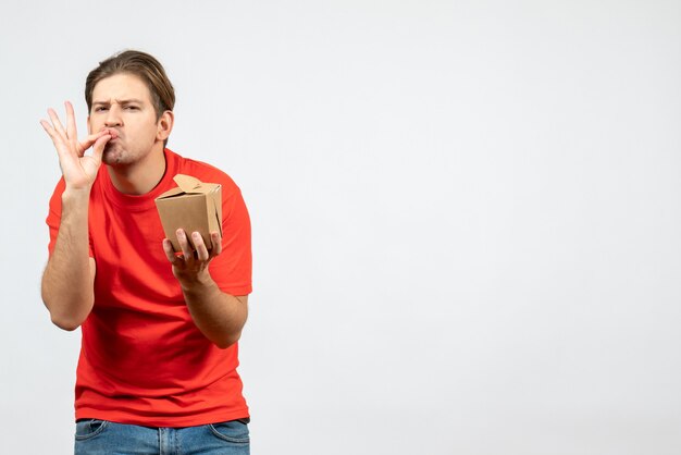 Vista frontal de um jovem confiante em uma blusa vermelha segurando uma pequena caixa, fazendo um gesto perfeito no fundo branco