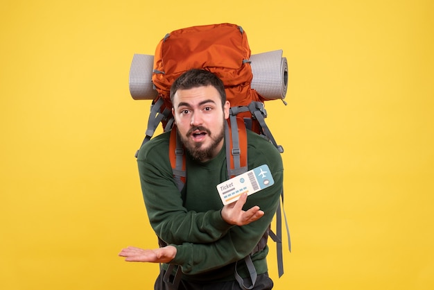 Vista frontal de um jovem confuso viajando com uma mochila e mostrando a passagem em fundo amarelo