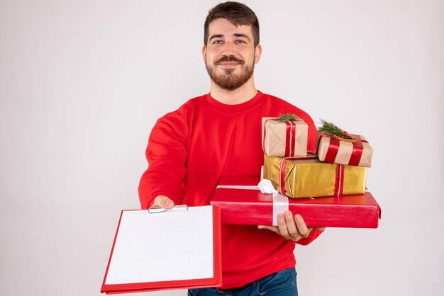 Vista frontal de um jovem de camisa vermelha segurando presentes de Natal na parede branca