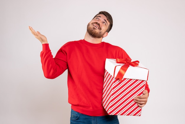 Vista frontal de um jovem de camisa vermelha segurando um presente de Natal em uma caixa na parede branca