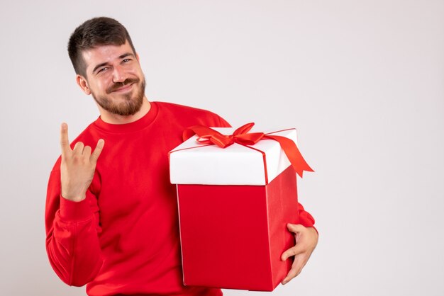 Vista frontal de um jovem de camisa vermelha segurando um presente de Natal em uma caixa na parede branca