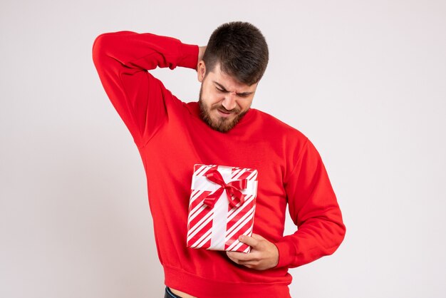 Vista frontal de um jovem de camisa vermelha segurando um presente de Natal na parede branca