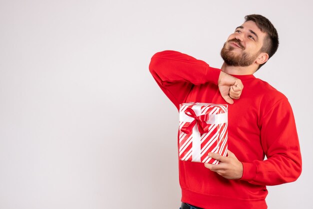 Vista frontal de um jovem de camisa vermelha segurando um presente de Natal na parede branca