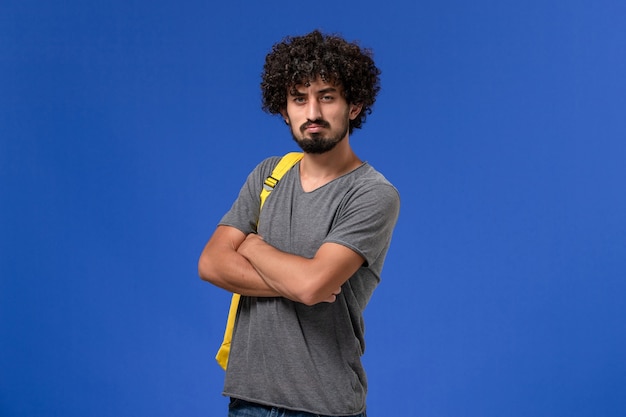 Vista frontal de um jovem do sexo masculino com uma camiseta cinza e uma mochila amarela, apenas posando na parede azul-clara