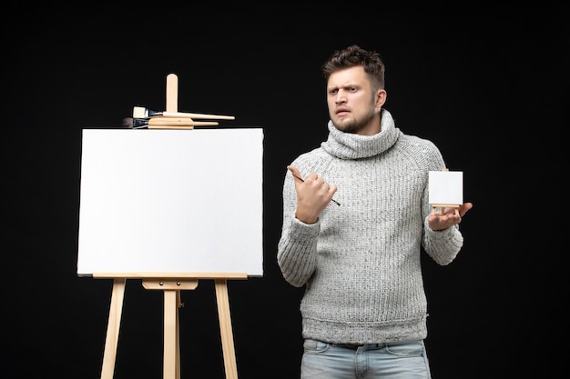 Vista frontal de um jovem e talentoso artista masculino inseguro, segurando o mini-livro com pincel e expressão facial de surpresa no preto