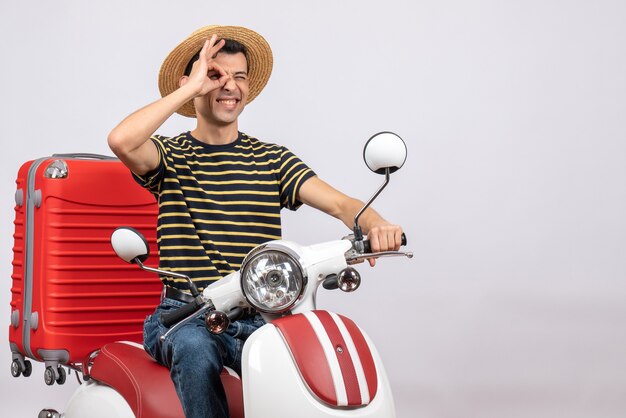 Vista frontal de um jovem feliz com chapéu de palha na motocicleta, colocando a placa de ok na frente do olho