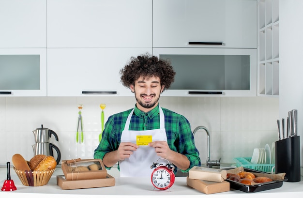 Vista frontal de um jovem feliz em pé atrás da mesa, vários bolos e segurando o cartão do banco na cozinha branca