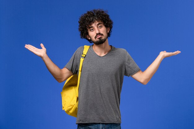 Vista frontal de um jovem homem com camiseta cinza e mochila amarela na parede azul