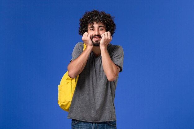 Vista frontal de um jovem homem com camiseta cinza e mochila amarela na parede azul