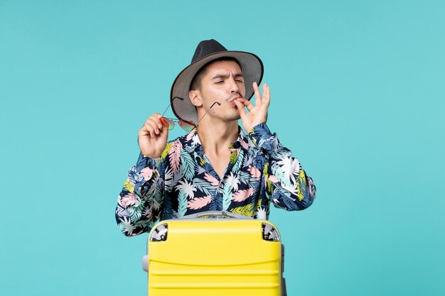 Vista frontal de um jovem homem com sua bolsa amarela se preparando para uma viagem, posando na parede azul clara