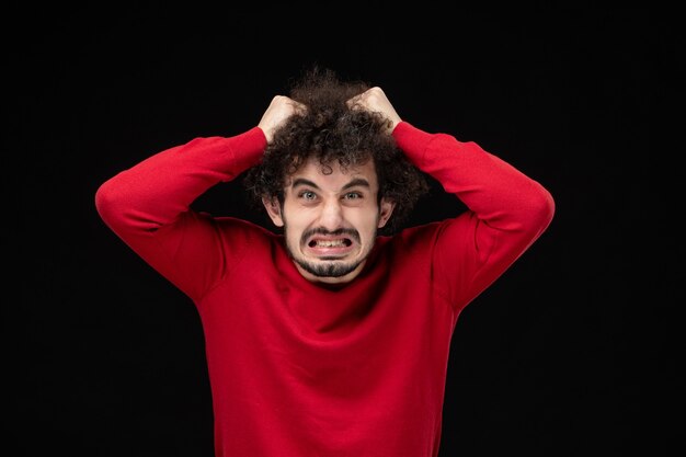 Vista frontal de um jovem homem de suéter vermelho, arrancando os cabelos na parede preta