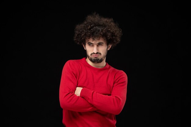 Vista frontal de um jovem homem de suéter vermelho com cara de louco na parede preta