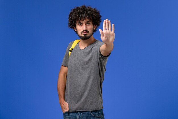 Vista frontal de um jovem homem em uma camiseta cinza usando uma mochila amarela mostrando o sinal de pare na parede azul-clara