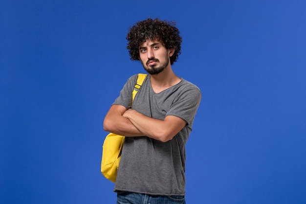 Vista frontal de um jovem homem em uma camiseta cinza usando uma mochila amarela, pensando profundamente na parede azul