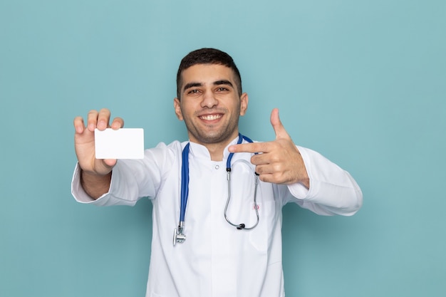 Vista frontal de um jovem médico de terno branco com estetoscópio azul segurando carddoctor branco